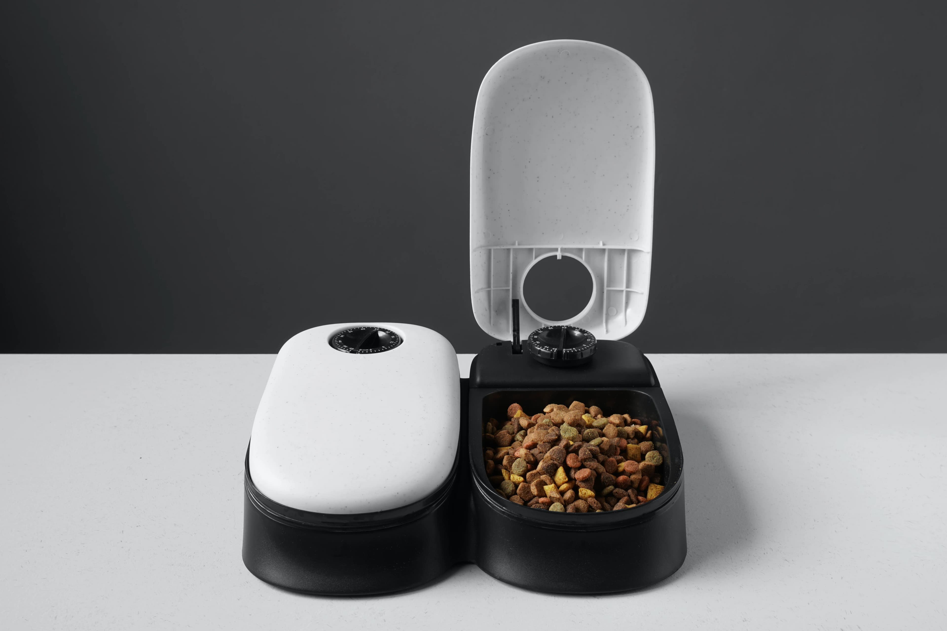 An automatic pet feeder with one lid open showing dry pet food inside and a timer dial, against a grey background.