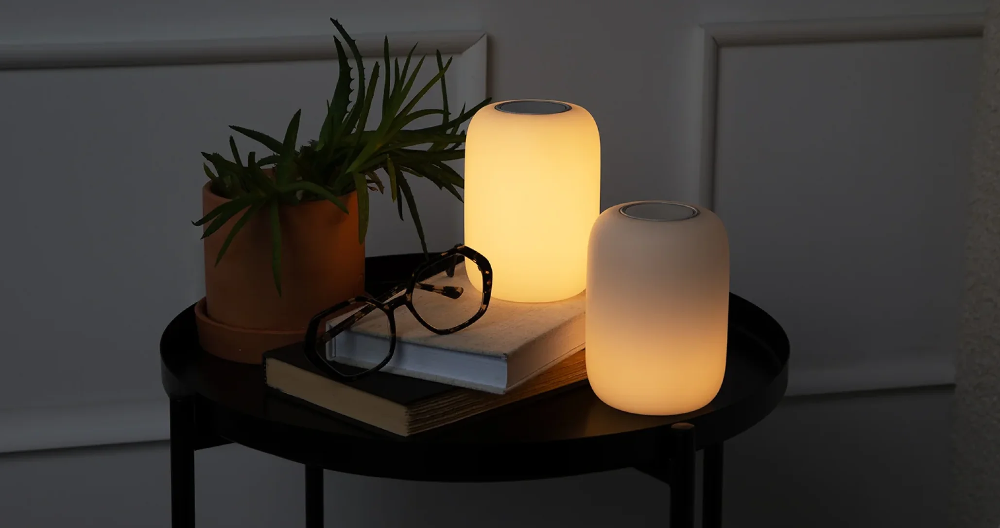 A cozy scene with two glowing candles on a round black table, next to a potted plant, a closed book, and glasses. The lighting is warm and soothing.