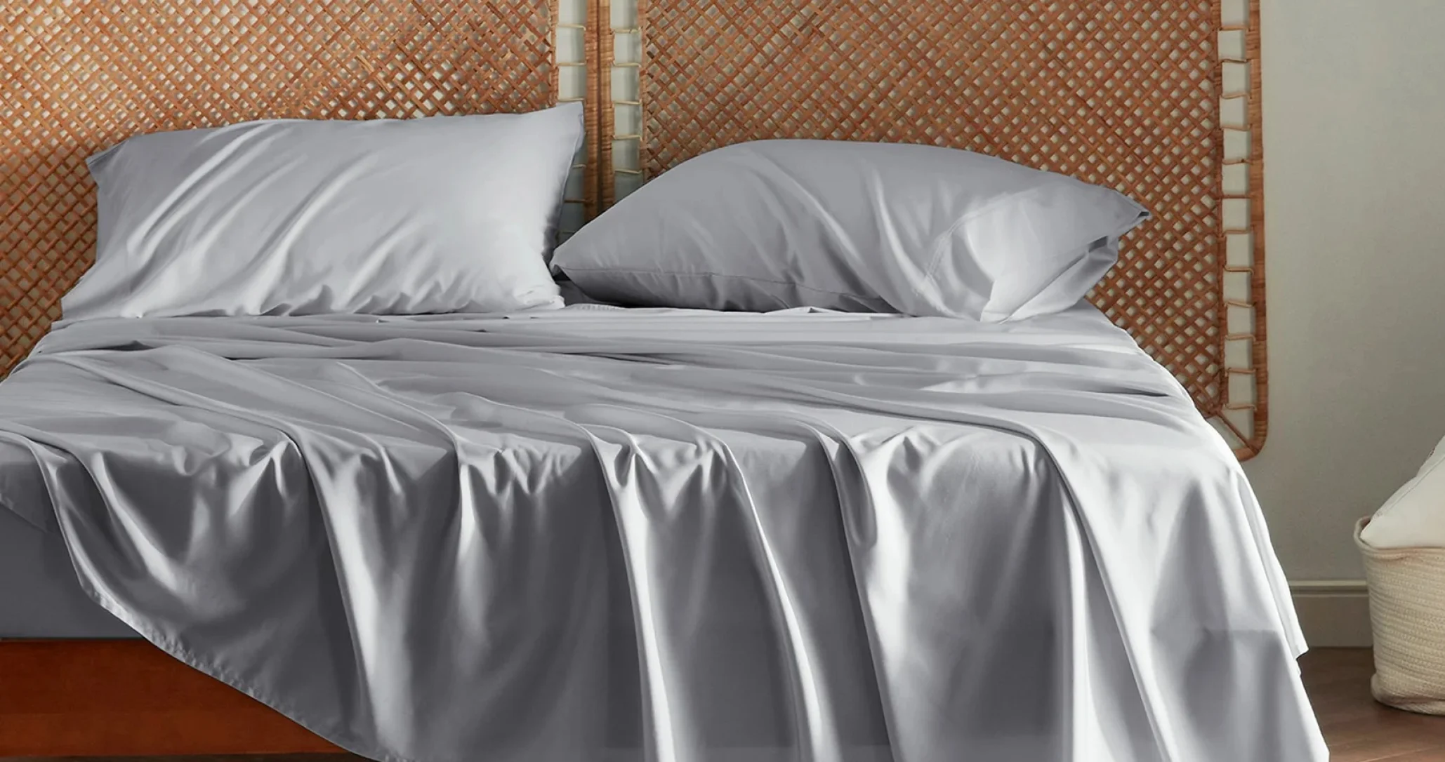 A neatly made bed with smooth, gray sheets and pillows, backed by a woven, earthy brown headboard. Soft, inviting, and serene bedroom scene.